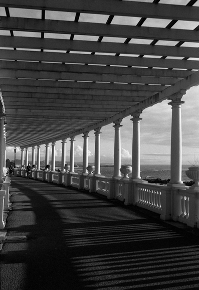 A monochrome image of a covered walkway with arches supported by white columns, leading towards an ocean view. Shadows create patterns on the path, and a few people are seen walking along the railing.