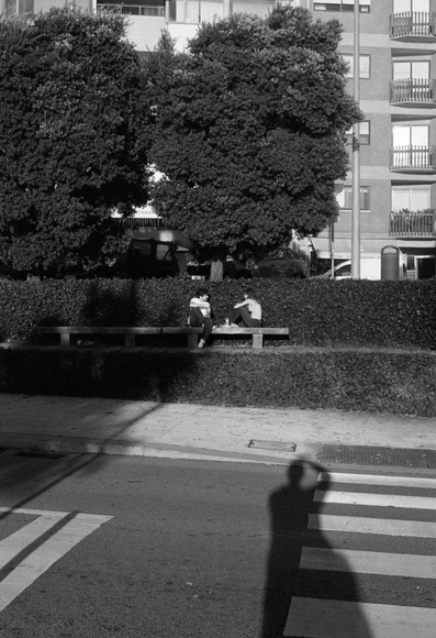 A black and white image featuring two people sitting on a bench surrounded by hedges and trees, with an apartment building in the background. An elongated shadow is cast on the road in front of them, indicating the presence of another figure nearby.
