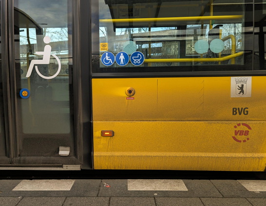 Blick auf ein Ausschnitt eines gelben Busses, unten graue Granitplatten, dahinter weiße breite Striche auf der Bordsteinkante, anschließend Bus. Links eine Tür mit Rollstuhl -emblem, in der Mitte ein Notöffnungsknopf. Darüber Fensterband. Tür und Fensterband in schwarz. Rechts Wappen von Berlin. Darunter BVG. Und VBB Log. Auf der Sonnegelben Seitenwand überall Drecksspritzer, die frontal von vorne nach hinten verlaufen. Notknopf ist halb dreckig und wirft einen "sauberen " Schatten in Richtung Tür.