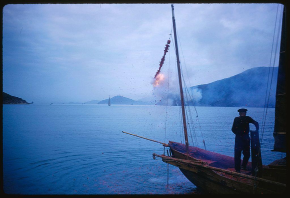 man on a sailboat in the harbor of hong kong, with fireworks going off behind him.