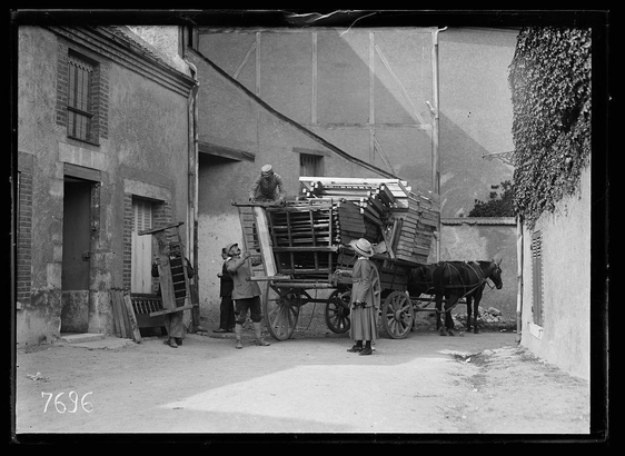 The image depicts a scene from what appears to be early 20th-century Europe, featuring several individuals engaged in the task of unloading and transporting furniture. A horse-drawn cart is central to the composition, with wooden crates piled on top, suggesting that these items were being delivered or moved.

There are at least four people visible: one man standing atop a ladder attached to the back of the wagon, seemingly securing or adjusting something; another person in mid-stride carrying what looks like two large pieces of furniture; and two more individuals conversing near the cart. The attire of these figures suggests modest social status, with simple coats and hats.

The setting is an urban alleyway lined by buildings exhibiting signs of wear and age, including overgrown vegetation on walls and a weathered facade on one structure to the left. A watermark or number "7695" appears in the bottom right corner, which could indicate cataloging information for historical records.

This image seems connected to a specific context involving refugees during World War I; it is attributed to Miss Morley of the American Red Cross Bureau of Refugees overseeing the distribution and sale of furniture made by French prisoners. The visual representation aligns with efforts documented in photographs taken by Lewis Hine, capturing various aspects of life during that tumultuous period [...]