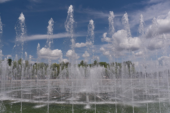 The image depicts a large fountain situated in the center of an open field. The fountain is filled with water shooting up into the air, creating a dynamic and lively scene. The sky above mirrors the fountain's activity by being blue and cloudy. In the background, there are trees that add depth to the landscape.