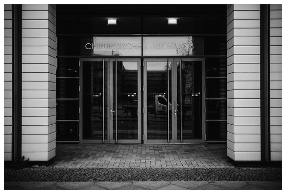 Frontaler Blick zwischen zwei Säulen hindurch auf eine Fensterfront mit Glastür. Über der Tür steht "Chirurgisches Klinik B" und auf den Scheiben "Sachsenklinik".