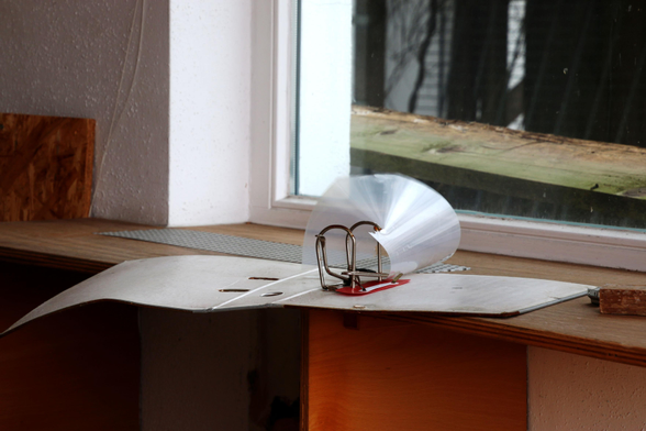 Ein aufgeschlagener Aktenordner mit einer aufgerollten Plastikschutzhülle liegt auf einer Fensterbank. Ein Stück Holz liegt draußen vor dem Fenster. Zu erkennen ist nur die Fensterecke in einem Abbruchhaus, weiße Raufasertapete, weißer Fensterrahmen, rotbraunes Holz für die Fensterbank. 

An open file folder with a rolled-up plastic protective cover lies on a window sill. A piece of wood lies outside the window. Only the corner of a window in a condemned building is visible: white woodchip wallpaper, white window frame, reddish-brown wood for the window sill. 