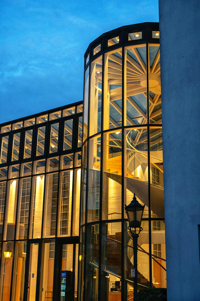 Das Bild zeigt ein modernes Gebäude mit einer markanten Glasfassade und einem zylindrischen, beleuchteten Treppenhaus.
Das Gebäude zeichnet sich durch eine Kombination aus Glas und dunklem Metallrahmen aus.
Im Inneren des zylindrischen Abschnitts ist eine Wendeltreppe sichtbar, die durch das warme Licht besonders hervorgehoben wird.
Die Szene spielt in der Dämmerung oder am frühen Abend, erkennbar am tiefblauen Himmel im Hintergrund.
Eine traditionelle Straßenlaterne im Vordergrund bildet einen interessanten Kontrast zur modernen Architektur.