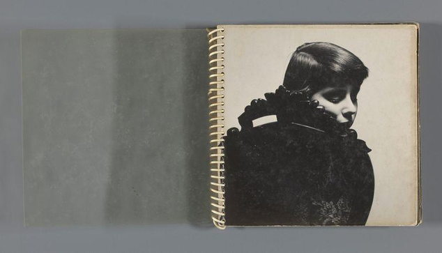 The image shows an open spiral-bound album or book with a single black and white photograph visible on the right page. The left side of the spread is blank, revealing only paper without any images or text. On the right page, there's a portrait-style photo depicting a young girl in vintage clothing, facing slightly to her right profile view towards us but not making eye contact directly with the camera. She has dark hair styled upwards and appears to be wearing heavy makeup around the eyes, including prominent eyebrows, eyeliner, and eyeshadow that gives an intense gaze or possibly somber expression.

The young girl is dressed in a black outfit adorned with what looks like ruffled fabric accents on her shoulders, suggesting it may be from a costume or theatrical performance. The background of the photograph is not visible as it cuts off at her chest level. The album itself has signs of wear and age, such as slight discoloration and some creases along the edges.

This image serves as an example for understanding what kind of content to describe if someone were unable to see it themselves, providing a detailed description based on visual elements present in the photograph without any text or caption.