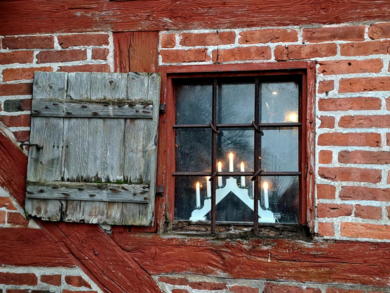Quadratisches Fenster in einer Fachwerkwand mit rotbraunen Balken und roten Ziegeln. Das Fenster ist durch rostige Metallrahmen in neun kleine Scheiben unterteilt. Links des Fensters ist ein hölzerner Fensterladen aufgeklappt. Hinter den Fensterscheiben steht ein kandelaber mit sieben elektrischen Kerzen.