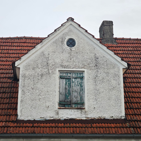 Das Foto zeigt eine Giebelgaube eines Hauses mit einem roten Ziegeldach. Die Gaube hat eine weiße, raue Putzfassade, die stellenweise verschmutzt oder verwittert aussieht. In der Mitte der Gaube befindet sich ein Fenster mit geschlossenen, verwitterten Holzläden mit abblätternder grüner Farbe. Oberhalb dieses Fensters ist ein kleines, rundes Fenster zu sehen. Rechts von der Gaube ragt ein Schornstein aus dem Dach. Mit dem Schornstein sieht die Gaube wie ein kleines Haus mit Schornstein aus.
Der Himmel ist bewölkt und hellgrau.