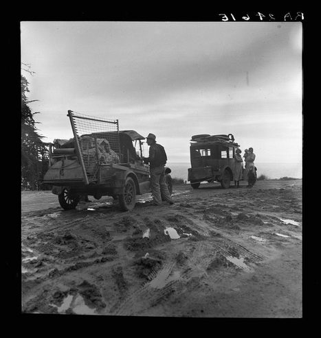 The image captures a moment of two families, possibly migrants from Missouri seeking work in the pea fields of California. This photograph is taken on an unpaved road where muddy tracks indicate recent rainfall or wet conditions.

In the foreground, we see one family standing beside their open-sided utility truck equipped with a cage for transporting goods. The man at center appears to be interacting with someone inside this vehicle, possibly communicating instructions or discussing travel plans. Another adult and two children are visible near him; they seem ready for an adventure ahead.

Parallel to them is another set of migrants traveling in what looks like a makeshift cart attached to a larger truck. This group consists of several adults, including women dressed in practical clothes suitable for fieldwork, alongside at least one child who appears eager or curious about their surroundings.

The background shows the sky and possibly early morning mist, giving off an atmosphere of hope mixed with uncertainty as these individuals embark on their journey towards work opportunities across states. The overall scene is evocative of a significant historical period when many families migrated in search of better living conditions during difficult economic times.