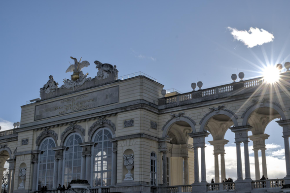 Das Bild zeigt ein beeindruckendes historisches Bauwerk. Es hat hohe Fenster und eine prächtige Fassade, die reich verziert ist. Oben auf dem Gebäude thront ein großer Adler. Die Architektur hat viele geschwungene Formen und Säulen.

Im Hintergrund ist der Himmel sichtbar, mit einigen kleinen Wolken und einem Sonnenstrahl, der durch die Wolken bricht. Diese Kombination von Licht und Schatten verleiht der Szene eine lebendige und dynamische Atmosphäre.

