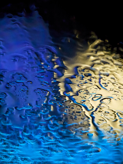 Abstract night-time image showing blue and yellow lights in the background to create an abstract pattern from rainwater pooling on glass. The manual focus is sharply on the rainwater on the glass surface, which makes the lights in the background distort, blend and flare into beautiful patterns.