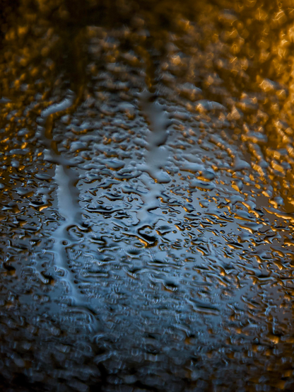 Abstract night-time image showing blue and amber lights in the background to create an abstract pattern from rainwater pooling on glass. The manual focus is sharply on the rainwater on the glass surface, which makes the lights in the background distort, blend and flare into beautiful and subtle patterns.
