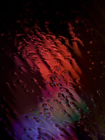 Abstract night-time image showing blood-red lights in the background to create an abstract pattern from rainwater pooling on glass. The manual focus is sharply on the rainwater on the glass surface, which makes the lights in the background distort, blend and flare into beautiful patterns.