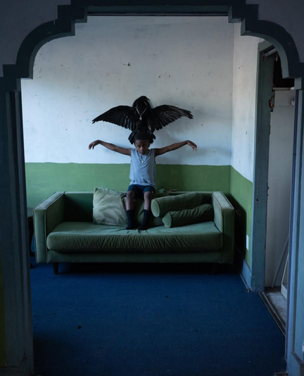 assis sous un corvidé empaillé cloué au mur, sur le dossier du canapé vert, un jeune garçon noir déploie ses bras comme les ailes de l'oiseau