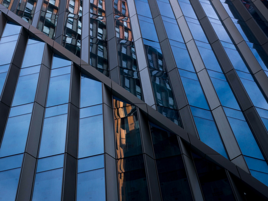 Photo of the glass front of a large building, the top-to-bottom windows corrugated like particularly angular ridged crisps. A diagonal frame slashes through the frame from top-left to bottom-right, dividing the fractured reflection of a many-windowed block and the sky above into two opposing triangles, the lower one with the sky to the left and the other with it at the right.