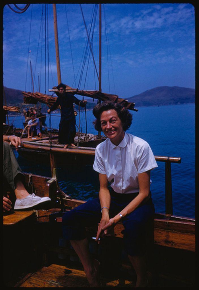 A woman is seated on a wooden bench on what appears to be an old-fashioned sailing ship, with her legs crossed and holding something in one hand. She has dark hair styled updo, wears glasses, white blouse tucked into navy blue knee-length skirt, silver bracelet on right wrist, watch or ring on left hand and holds cigarette between fingers of same hand which rests near the bench's edge.

In the background we see a man wearing dark clothes at ship's stern handling what could be rigging. Another person is barely visible to his right side sitting down with legs crossed as well. The sea stretches out behind them, reflecting light from clear blue skies and hinting that it might not actually be sunny since the water looks calm. In the far background we can see a landmass or hillside.

The photo carries an inscription: "Sightseeing in Hong Kong / July 1959" along with information about photographer Toni Frissell, associated with Sports Illustrated magazine and her work being part of this assignment to capture scenes from Hong Kong. The photograph is noted as #X5826 (Extra) taken by the International Correspondence News Service.

This image captures a moment likely meant for leisure or tourism, showcasing an older style boat commonly used in historical times rather than modern day cruising vessels which are typically larger and with amenities such as restaurants and entertainment  [...]