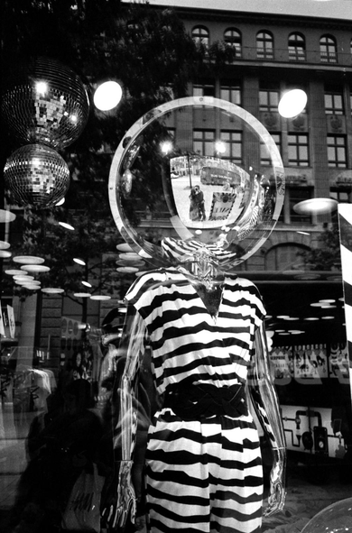 black and white portrait photograph of a mannequin in a shop window. the mannequin has a perspex sphere as a head. the photographer and his partner are visible reflected in the headspace.