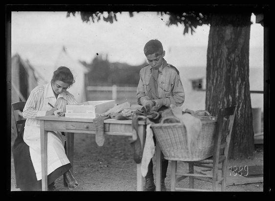 The image shows two individuals, possibly during World War I era based on their attire and the setting. The person on the left appears to be a woman in civilian clothing with glasses, writing or examining something at a wooden table laden with various items including papers, boxes, and what seems like medical supplies such as gloves.

The individual on the right is wearing a military uniform similar to that of an army soldier from around World War I. This person stands beside another outdoor setup where there's a large wicker basket filled with folded clothing or blankets next to a bench. A tree trunk prominently frames the scene, suggesting they are in some sort of field hospital camp near Paris.

The overall atmosphere is one of diligence and duty as both individuals engage in tasks that seem related to patient care or military organization. The black-and-white nature of the photo suggests it's an old photograph capturing a moment from history.