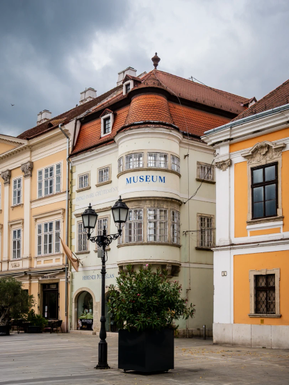 Städtischer Bereich, Fußgängerzone. Drei Wohnhäuser unterschiedlichster Art  nebeneinander: mit rundem Erkerbereich, klassizistisch, Gründerzeit. Alle zeigen ihre unterschiedlichen Fensterfronten.
Davor ein großer Blumenkübel und eine Doppellaterne.