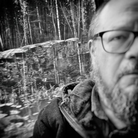 A black-and-white photo capturing a passing landscape through a train window with trees and rock formation and a person in the foreground.