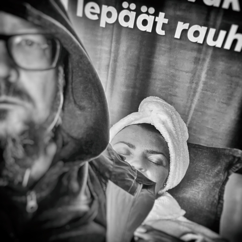 A man with a beard in a hooded jacket poses in front of a backdrop with Finnish text, creating a moody black and white atmosphere.