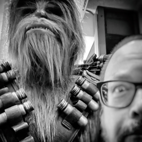 A close-up of a Chewbacca costume with a surprised man in glasses in the foreground, captured in black and white.