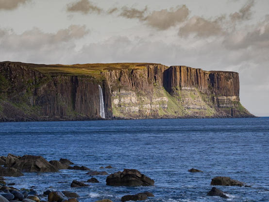 A view across the sea from a rocky foreshore across a bay to some columnar cliffs with a spectacular waterfall cashing down them.
