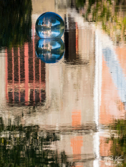 Photographie en couleur de format vertical, présentant dans son intégralité, un reflet légèrement troublé capté à la surface d'un bassin.
L'objet du reflet est une partie d du pignon d'un château constitué de pierres et de briques avec notamment, une haute baie vitrée peinte en ocre, à peu près comme les briques du mur, et une porte ouverte.
Sans doute que le sujet le plus intéressant de la photo est une sphère remplie d'eau installée juste à la surface de l'eau du bassin et juste en dessous son propre reflet. Dans la sphère, on peut voir l'intégralité du château et une grande partie du bassin avec par conséquent, le reflet du château ce qui fait  2 châteaux + 2 châteaux du reflet de la sphère… 4 châteaux en tout. ! 
Et en prime, un ciel magnifiquement bleu et son reflet multiplié par 4 !
