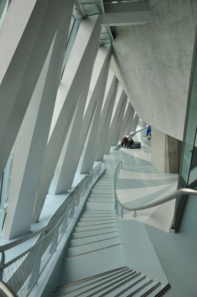 Durch ein winklig angeordnetes Spalier von Betonpfeilern fällt diffuses Licht von der linken Seite ins Treppenhaus des Daimler Museums in Stuttgart. Die gesamte Architektur ist bis auf das Edelstahlgeländer in weiss gehalten und unterstreicht damit die Tiefe des dargestellten Raumes.