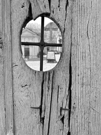 The photo is in black and white. shows a weathered wooden gate with a circular window, reinforced by crossed metal bars, reveals a view of a graveyard.