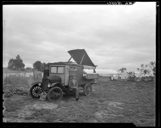 The image is a black and white photograph depicting an old-fashioned pickup truck parked on what appears to be rural farmland. The vehicle, with its back gate open and wooden bed, suggests it was used for transporting goods or materials over long distances during times when modern transportation methods were less prevalent.

A man stands beside the truck, leaning casually against its side. He is wearing a suit jacket, trousers, hat, and appears to be engaged in conversation or observing his surroundings with contemplation.

The environment includes an open field strewn with debris like wood planks, suggesting recent activity that may have involved farming work. In the background, there are makeshift structures such as tents made of tarpaulin, indicating a possible temporary settlement for agricultural laborers. Clotheslines strung across show laundry drying in the air.

The photograph is marked "Dorothea Lange" and has annotations including "Yankee Doodle," "Nipomo CA," along with some unclear handwriting which may reference additional context or identifiers about this particular scene from One pea picker's home, located off Highway 101 at Nipomo, California. This image likely documents the everyday life of agricultural workers during a period when manual labor was more common in farming communities.

The photograph carries historical significance as it captures an era an [...]