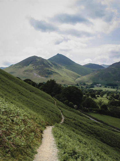 A winding path (left) leads across the side of a hill above a wooded valley. In the background are a series of peaks under a partly cloudy sky.