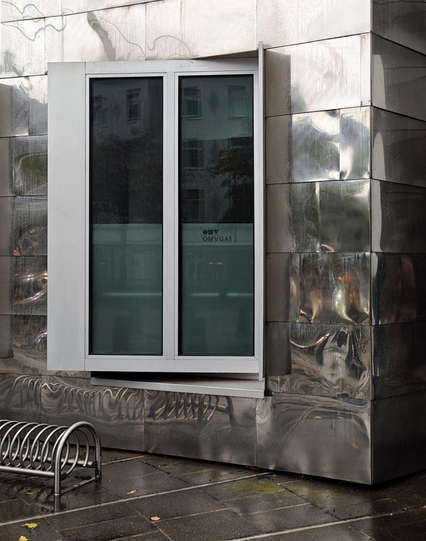 A photo of a reflective metal building facade with a large window with a white frame. A small metal bike rack stands on a wet sidewalk. Everything is shiny excited for the white window frame.

