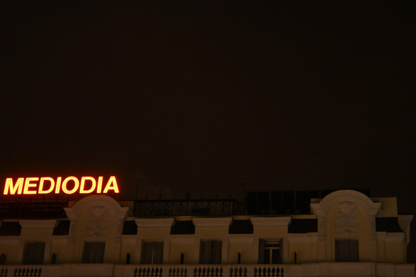 Fotografía nocturna; se muestra el extremo superior de un edificio con un cartel luminoso de neón que dice "MEDIODIA". Las tres cuartas partes superiores de la fotografía las ocupan el cielo nocturno despejado.