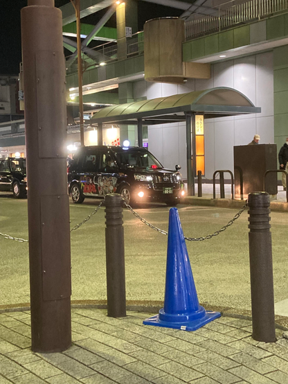 Image shows an urban street at night, featuring a blue traffic cone in the foreground, with a metal post and chain nearby. In the background, a person is pushing a stroller, while another person walks away. Storefronts with lights are visible.