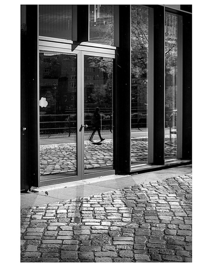 A high-contrast black and white photograph captures a modern building facade and the cobblestone ground in front of it. In the foreground, the textured, uneven stones of the pavement are emphasized by strong sunlight and deep shadows. Above the pavement, the building features large vertical glass panels framed by dark metal. Reflected clearly in the central glass panel is the silhouette of a person walking from right to left, carrying a bag. This reflection also shows the wet cobblestones where the person walks, along with the bare trees and a building across the way. The sunlight casts defined, sharp shadows on the ground and frames the scene.