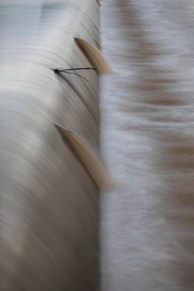 Across a river is a weir. A smooth concrete dam only used when it rains hard. It rained hard so there is water flowing over the perfectly straight man made dam. The water is gray and brown. There are three things breaking up the perfectly smooth long exposure flow. Two minor flaws in the dam and a branch stuck at the top. Beautifully unnaturally smooth flow gray perfect when things should not be perfect. 