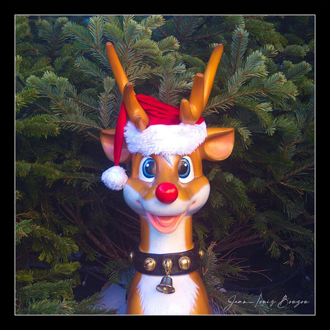 A cheerful, cartoonish reindeer bust fills the frame, its oversized blue eyes wide and glossy with bright catchlights that make it look almost alive. A perfect little red nose sits front and center — shiny and round like Rudolph’s — and a jaunty Santa hat, plush white trim and pom-pom included, is cocked to one side atop the reindeer’s golden antlers. The face is painted with warm, creamy tones and a broad, friendly grin exposing a hint of tongue, giving the figure an innocent, playful expression. Around its neck hangs a black collar studded with round brass bells and a slightly larger bell that dangles at the throat, promising the soft jingle of holiday cheer.
Behind the figure, dense, deep-green fir branches create a rich, textured backdrop that contrasts with the glossy, smooth surface of the reindeer. Warm lighting skims across the plastic-like finish, accentuating highlights on the nose, antlers, and the rim of the hat while casting soft shadows into the pine boughs. The overall effect is festive and nostalgic — a whimsical holiday ornament frozen in a moment of bright, unapologetic joy.

