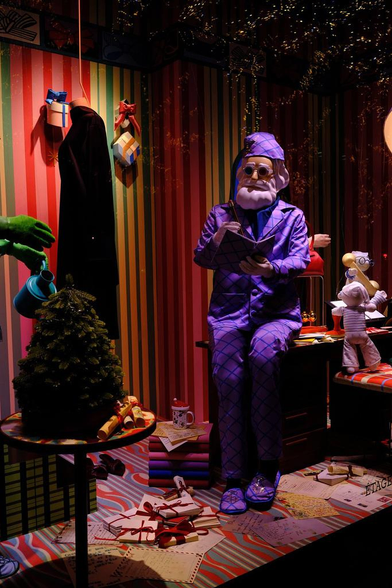 Photo d’une vitrine de Noël avec un père noël assis sur un bureau, en pyjama violet (avec le bonnet) et des lunettes de soleil rondes, faisant un peu penser à John Lennon