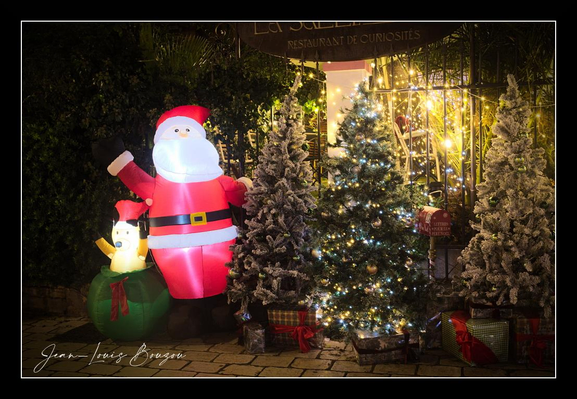 The scene is a small, festive tableau lit against the dark of night. On the left, a plump inflatable Santa glows from within — his red suit and white beard gently illuminated, one arm raised in a friendly wave. At his feet a cheerful inflatable penguin wearing a red hat sits atop a green gift, adding a playful, childlike touch.
Center stage is a medium-sized Christmas tree strung with warm white lights that twinkle softly, its branches dusted with artificial snow. Flanking the main tree are two smaller, heavily snow-frosted firs, creating a miniature snowy grove. Wrapped presents in red, green and metallic paper are scattered around the bases of the trees, and a small red sled and other seasonal props add to the tableau.
Behind the display a curtain of warm fairy lights and dark foliage provides a glowing backdrop, producing a cozy halo effect and gentle reflections on the stone-tiled ground. The overall impression is welcoming and whimsical — a compact, well-lit holiday vignette that mixes traditional decorations with a playful, modern touch.

