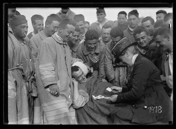 A black and white historical photograph depicting a group of individuals, likely convalescents or patients in a military hospital setting. In the foreground is an individual lying down with bandages on their arm and leg, receiving attention from another person seated beside them who appears to be handing over money cards as tokens for encouragement or reward. The surrounding crowd includes people wearing uniform-like garments suggesting they may be soldiers or workers associated with the institution. Many in the group are smiling or laughing, indicating a jovial atmosphere despite what seems like challenging circumstances.

The image is marked by date stamps at various locations (1928 and 17/15), potentially referencing specific events within the historical context of World War I-era France where such convalescent hospitals were active. The text overlay from an accompanying source suggests that these individuals are being comforted or given tokens to boost their morale, highlighting a moment of humanity amidst adversity during wartime recovery efforts.

Additional information about this image can be found in "Your hard luck is past," which seems like the title or heading for the photograph along with some descriptive narrative. The link provided points towards further details and context on an archive website dedicated to historical photographs by Lewis Hine, a renowned p [...]
