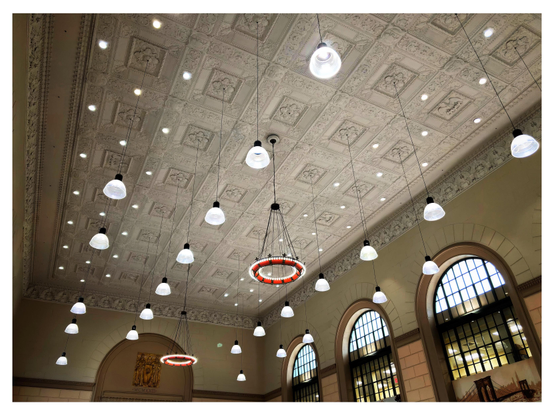 Photo of a majestic building interior, looking upwards. Tall arched windows sit under a high ornamented ceiling with a variety of different lights: some embedded in the ceiling surface, some hanging down in half-shell casings suspended from cables, and two antique chandeliers, each consisting of a reddish ornamented metal hoop ringed by small lights. On the far wall, under an arched design echoing the windows, is a metal plaque with bas-relief figures that reads “Justice” and “Prudence” with “MCMXXIII” underneath (1923, the year that ground was broken for the building).