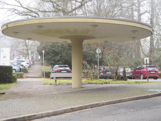 Auf einem stabilen runden Betonpfosten ruht ein kreisrundes ausladendes Dach als Überdachung der Bushaltestelle auf dem Halberg. Unter dem Dach sind runde Lampen angebracht und im hinteren Teil steht eine Parkbank.