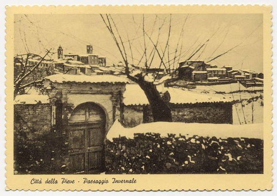 The image displays a postage stamp featuring an old, sepia-toned photograph of Città della Pieve during winter. The landscape captures snow-covered rooftops and bare trees against a pale sky in the background. Prominent architectural elements include arched gateways with doors, suggesting historical significance. A cluster of buildings can be seen rising behind these structures, creating a layered visual effect indicative of an old town setting. No discernible movement or people are present, emphasizing stillness. The text on the stamp reads "Città della Pieve - Paesaggio Invernale," indicating that it is showcasing the winter scenery of Città della Pieve.