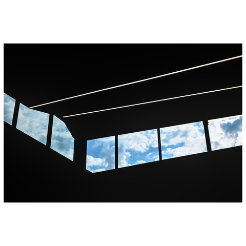 This image shows a segmented window front beneath a dark ceiling. The windows frame views of a bright, partly cloudy sky. The window segments are aligned diagonally, creating a sense of movement and perspective. Two led light strips on the ceiling add two strong lines to the composition. The contrast between the dark interior and the bright sky outside is sharp and distinct. The clouds in each skylight appear soft and fluffy. (drafted with Mistral.ai, edited by author)