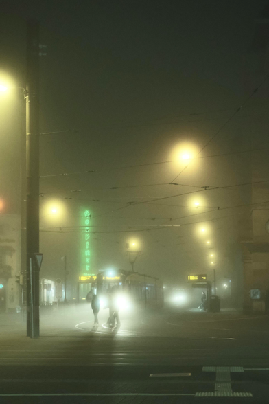 A photo down a street with a street level metro station. There are cables in the dark sky, illuminated by many bright lamp spots. The train itself has its headlights turned on, their rays are broken by three individuals walking by. It’s a glowy scene 