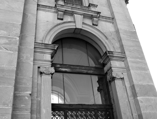 A black and white photograph taken at an angle, looking upwards, of an arched window adorned with Corinthian columns in a rectangular stone wall. The building is a kind of square tower with windows on three sides. Another similar window is therefore visible through it, as well as the ceiling decoration inside. Another, smaller window is partially visible above.

Photographie noir et blanc prise en angle en regardant vers le haut, d'une fenêtre en arche ornée de colonnes à chapiteaux corinthiens dans un mur en pierres rectangulaires. Le bâtiment est une sorte de tour carrée avec des fenêtres sur trois côtés. Une autre fenêtre semblable est donc visible au travers, ainsi que la décoration du plafond à l'intérieur. Une autre fenêtre plus petite est en partie visible au-dessus. 