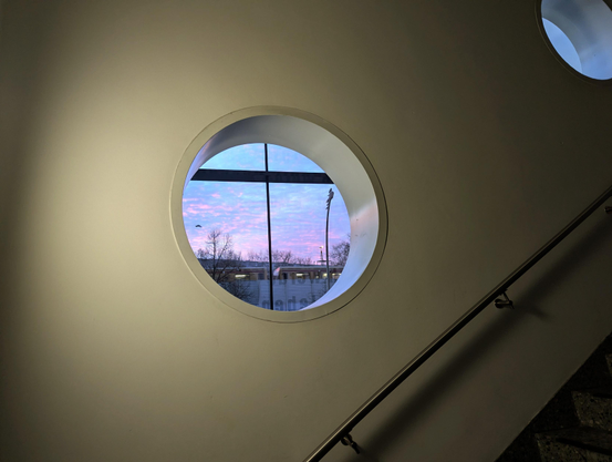 Weiße Wand mit Lichtkegel und zwei runde Fenster. Eins mittig, Mi Blick auf bunten Himmel, vorbeifahrende S-Bahn und auf Laterne sitzende Tauben. Rechts Ausschnitt des Handlaufs, ohne schmuck. Granittreppenstufen in rechter unteren Bildecke 