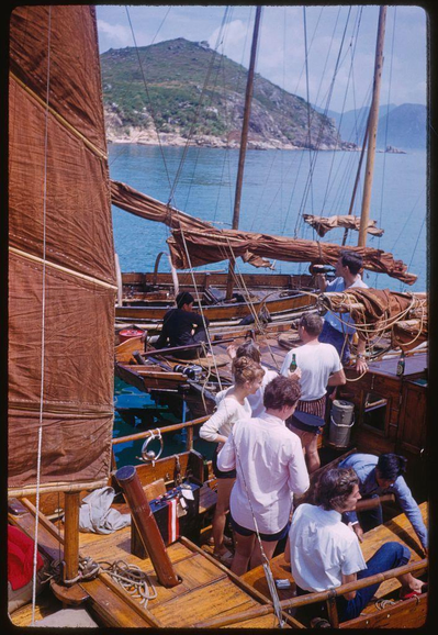 A group of people on a wooden boat with tall, brown sails. The setting appears to be in the open water near an island or large hillside. Several individuals are casually dressed and socializing while one man looks through binoculars at something off-camera. There is visible equipment including buckets and what seems like a small portable generator or battery pack. One of the men has his arm around a woman's waist, suggesting intimacy between them. The overall mood appears relaxed as people unwind on their boat during leisure time.