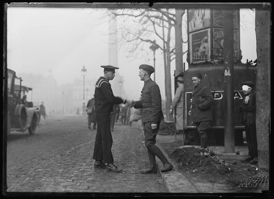 The image captures a moment on an urban street, possibly during or just after World War I. Two men in military uniforms are engaged in conversation with each other; one is dressed as a sailor while the other wears army attire. Behind them stands another man in civilian clothing observing their interaction.

In the background, there's a bustling cityscape filled with pedestrians and vehicles typical of early 20th-century urban life. A vintage car, likely from the same era depicted by the uniforms, occupies part of the street alongside an old bus or trolley labeled "Paris." The atmosphere appears to be one of quiet conversation amidst daily activities.

The image has a historical context, suggesting it was taken during a time when soldiers were stationed in Paris as part of Allied forces. It encapsulates interactions between military personnel and civilians at that momentous period in history.
